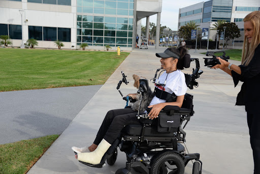 A woman is sitting in a black motorized wheelchair and consuming information on a handled device,